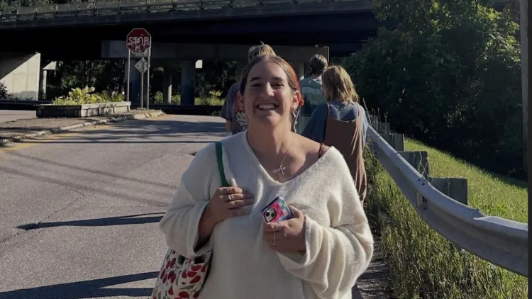 Person standing outdoors with a bridge and greenery behind.