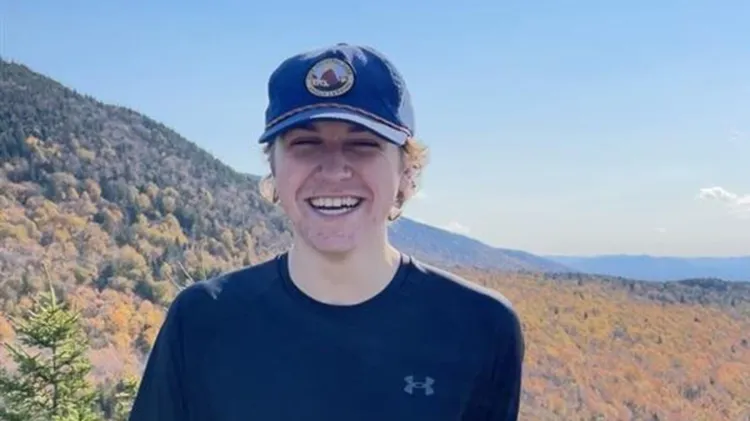 Person smiling outdoors with mountain and fall foliage in the background, wearing a black shirt and blue cap.