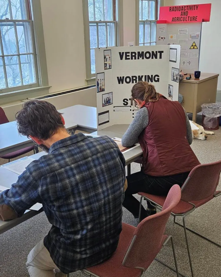 Two people are seen from the back, sitting at a table in front of posterboards with titles like "Vermont Working Steer" and "Radioactivity and Agriculture"