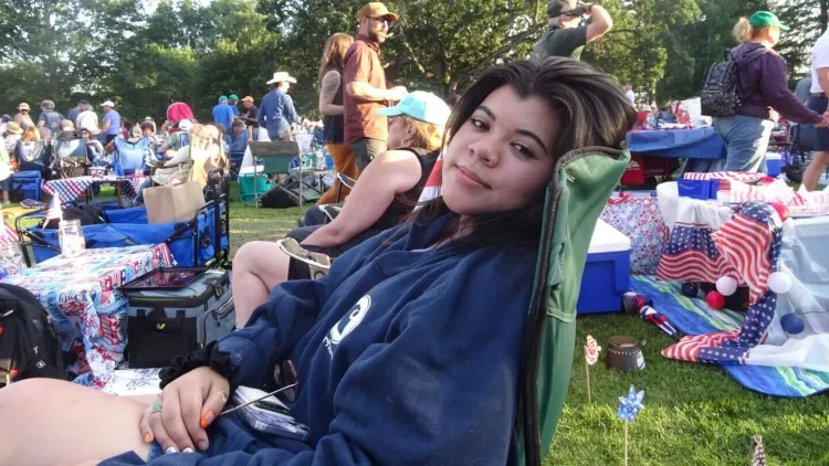 Person relaxing in a lawn chair at an outdoor gathering with a crowd and picnic setup in the background.