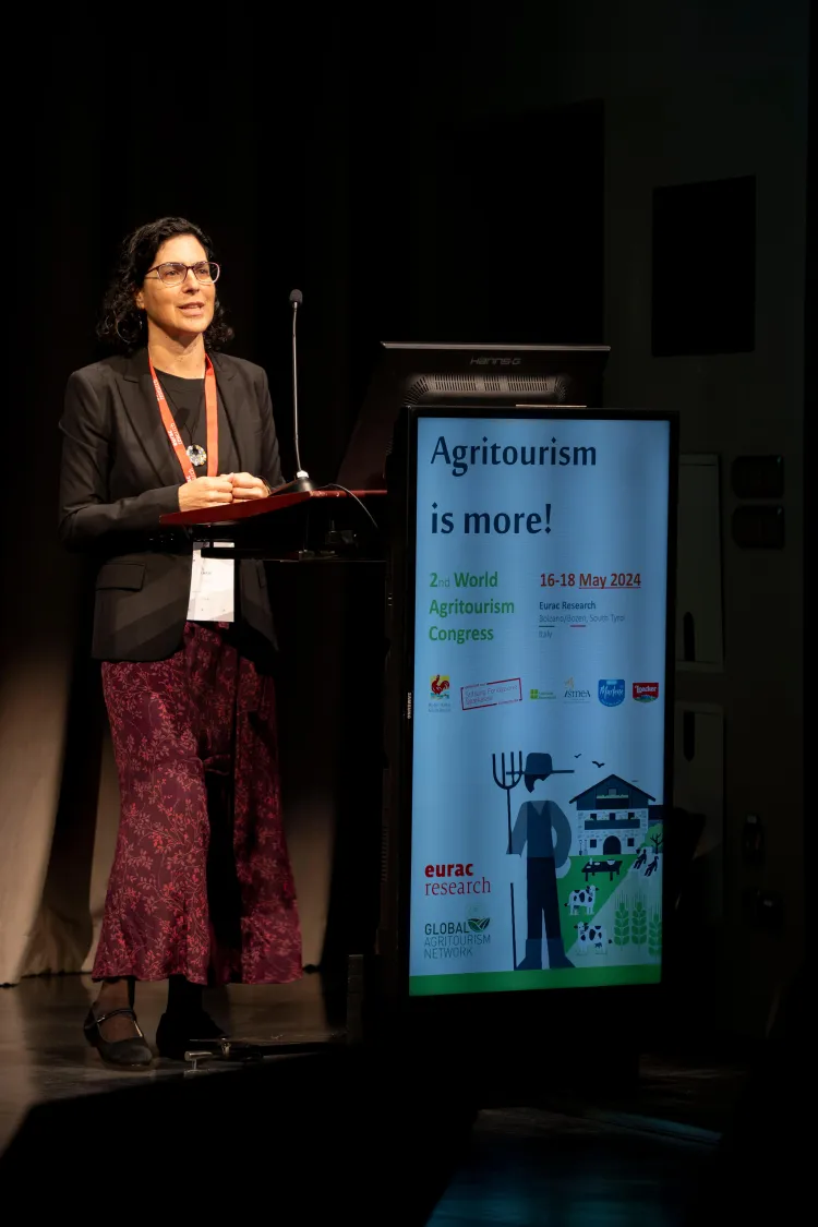 A person at a podium speaking and standing by a screen reading "agritourism is more!"
