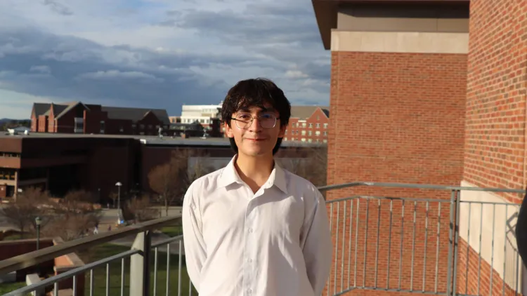 Person standing on a balcony with campus buildings and a cloudy sky in the background.