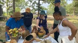 people stand around a table outside. there are flowers and various containers on the table