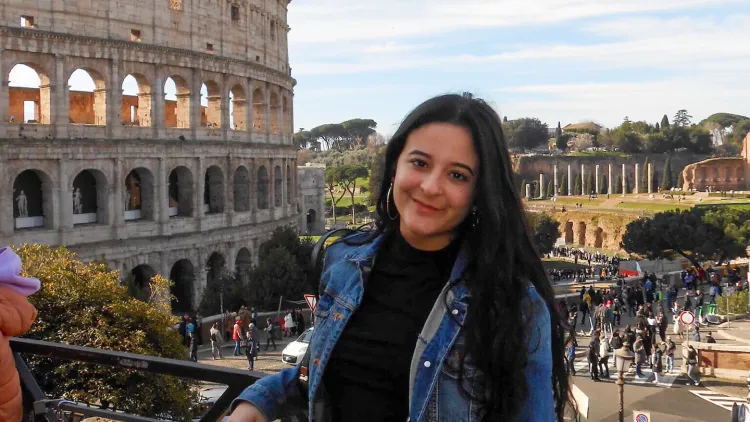 Person posing in front of the Colosseum with a crowd and historic buildings in the background.