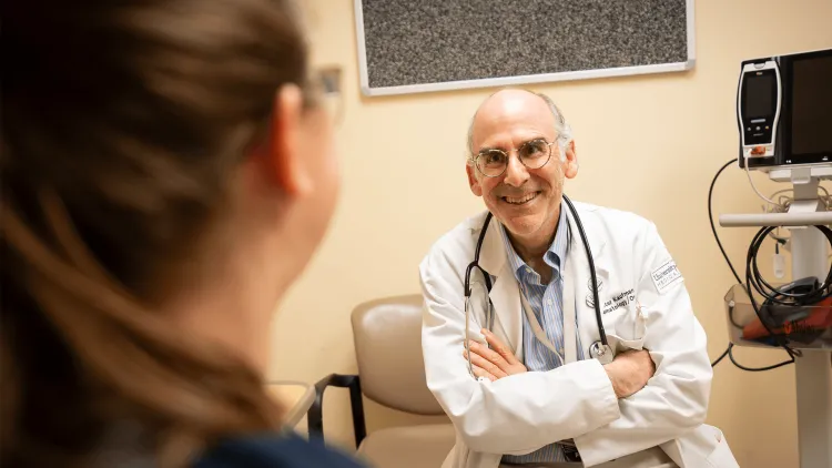 A doctor in a lab coat with arms crossed talks with a patient. 