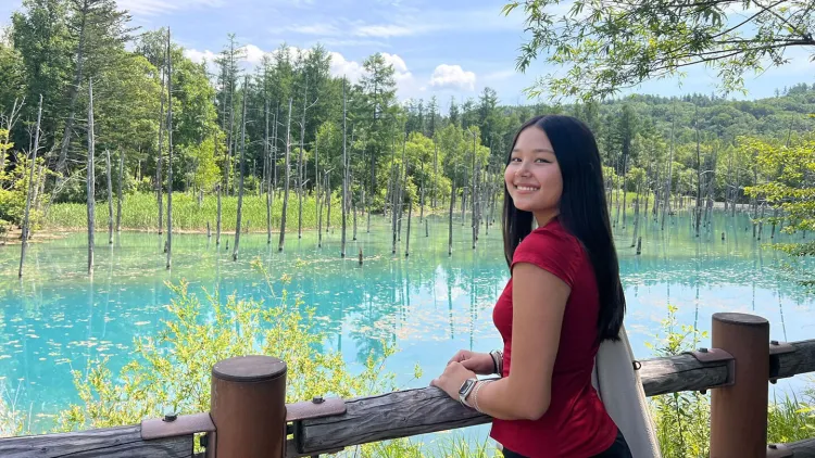 Person standing by a railing next to a bright blue lake with trees in the background.