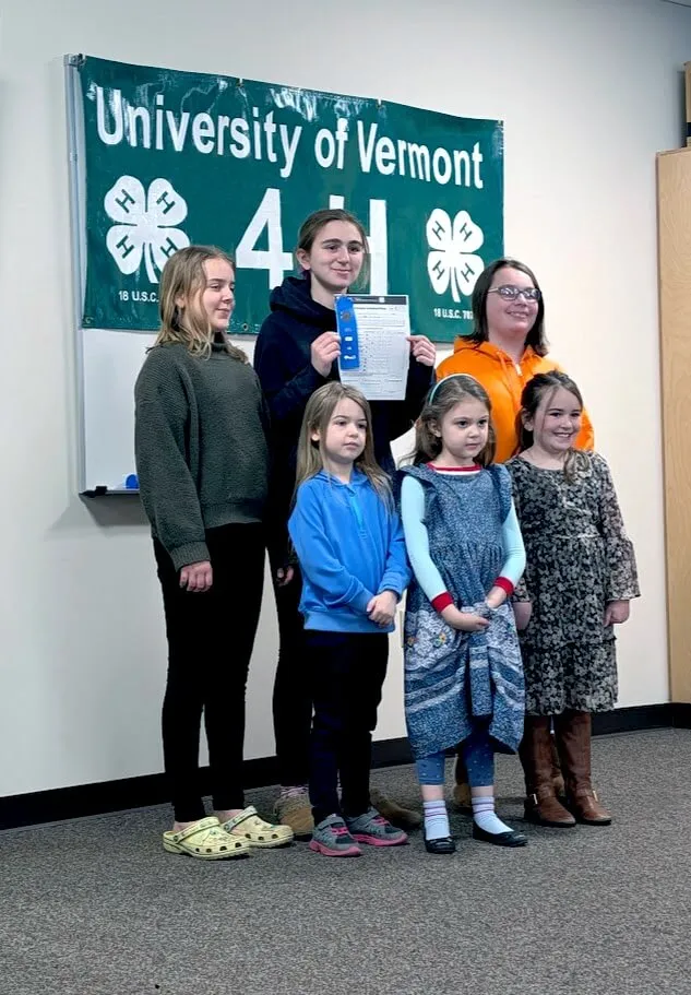 A group of six youth stand in front of a green poster reading University of Vermont 4-H with clover logos.
