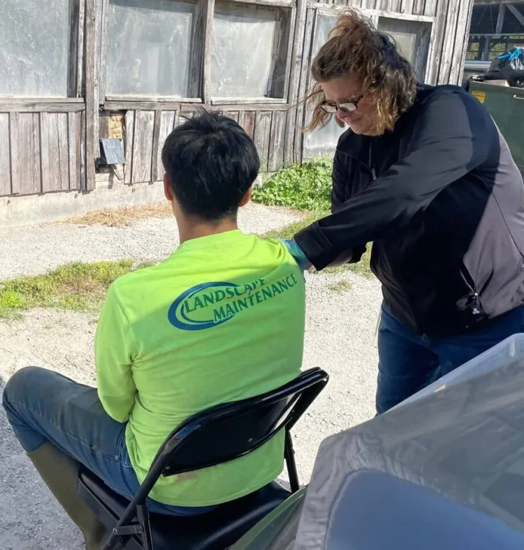 A photo of a person in glasses administering a vaccine to a person sitting in a chair, seen from the back and wearing a landscaping shirt and muck boots