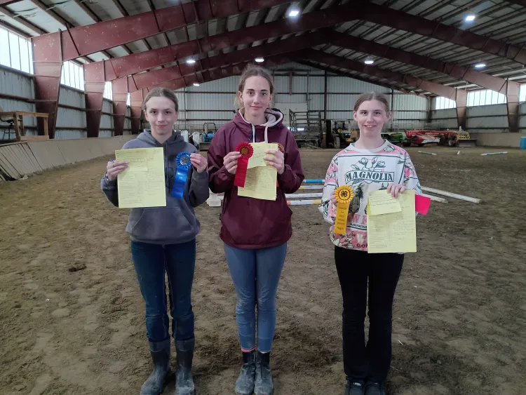Three young people stand holding rosette ribbons inside an indoor horse arena, with sand on the ground and a long open space.