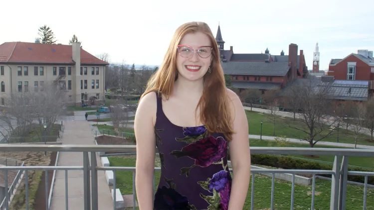 Young woman with long red hair and glasses, in dark flowered dress, standing by railing with campus in background