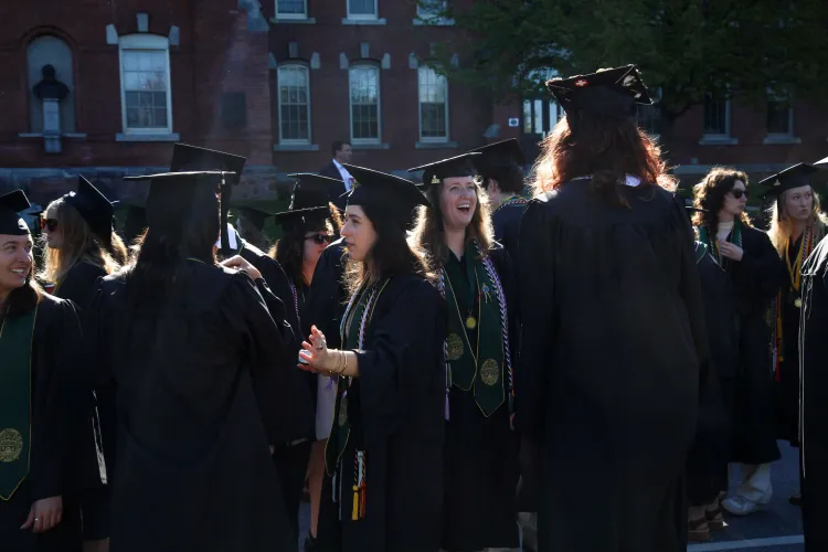 Young people in caps and gowns are backlit by the sun, and a brick building is behind them.