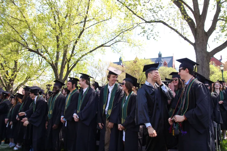 People in caps and gowns laugh and stand together under tall green trees.