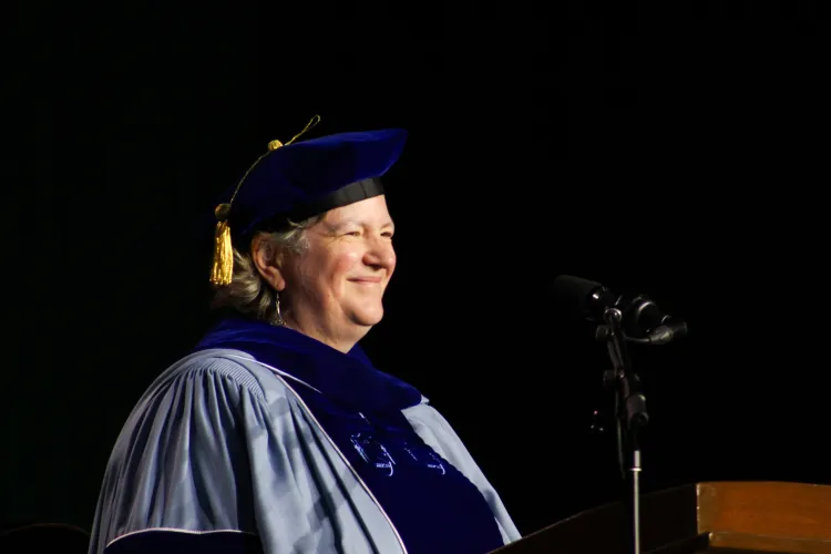 A person in velvet blue cap and gown smiles at a microphone.