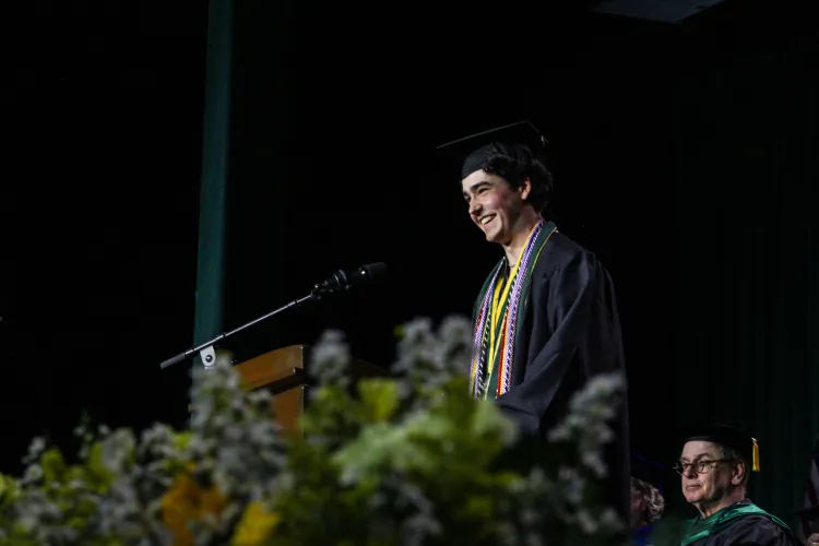 A young person smiles at a microphone, wearing cap and gown. Flowers are in the forefront.