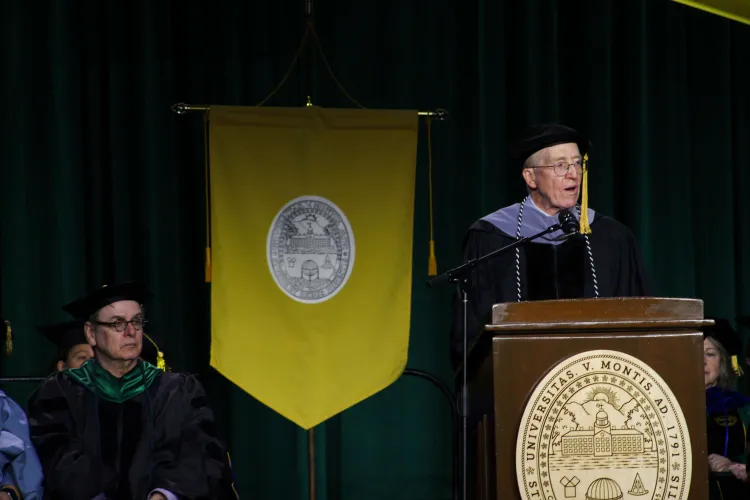 A person in a cap and gown speaks at a podium, with a yellow flag in the background.