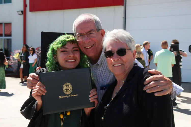 Three people hug. A younger person with green hair holds up a diploma while two older people lean in, and all smile.