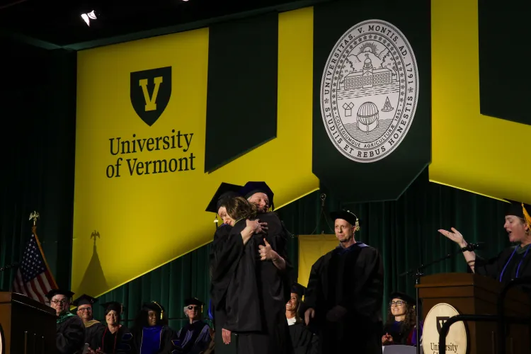 Two people in caps and gowns hug on stage with a giant yellow and green UVM banner in the background.