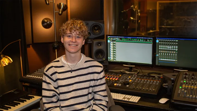 Young man with curly blond hair and striped shirt sitting in front of sound board in studio