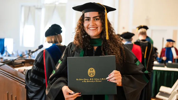 a person wearing regalia holding a diploma with other people wearing regalia in the background