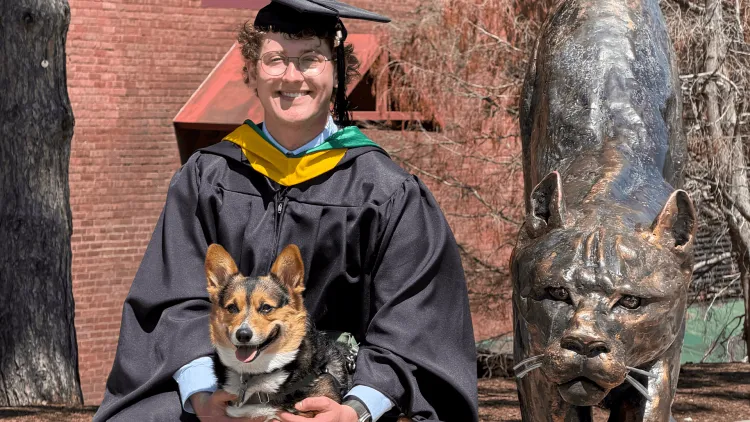 a person in academic regalia with a dog, next to a catamount sculpture