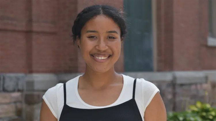 Young woman with dark hair, wearing white and black top, smiling and standing in front of brick building