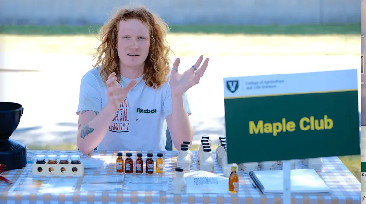 A person sitting at a table with maple syrup samples and a sign that reads Maple Club