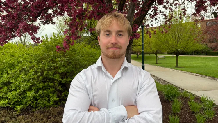Young man with blond hair and cropped beard wearing white shirt and standing on campus green