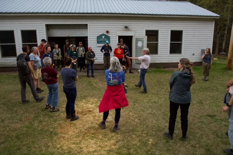 A couple dozen people stand on grass outside a white building. A woman in a pink shirt and jeans points in the middle of the group