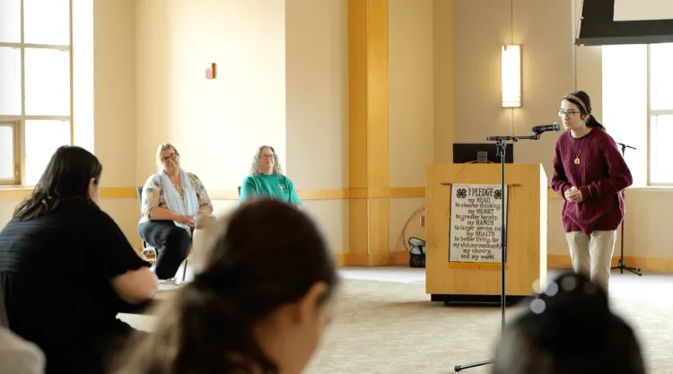 A young person speaking into a microphone, with audience members visible in the foreground.