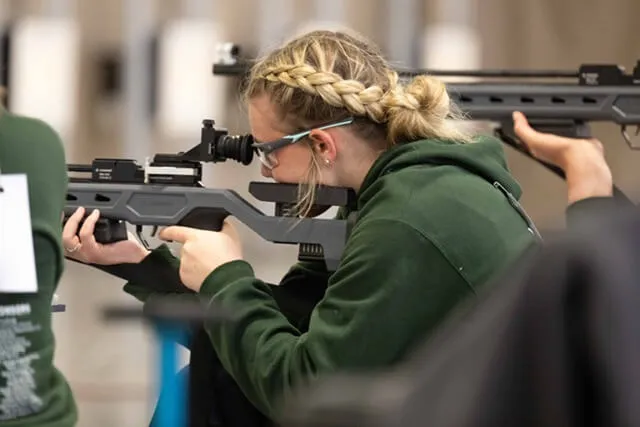 A young person in a green sweatshirt and glasses, with blonde french-braided hair, looking into the scope of an air rifle. More young people holding rifles are in the foreground and background.