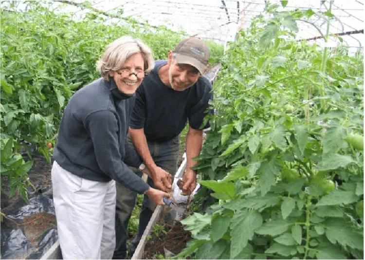 Two people pose for a photo inside a hoophouse with tall plants.