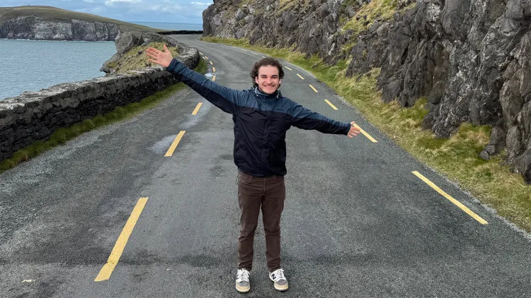 Smiling young man in blue jacket with arms outspread standing in middle of road with mountain on one side and ocean bay on the other