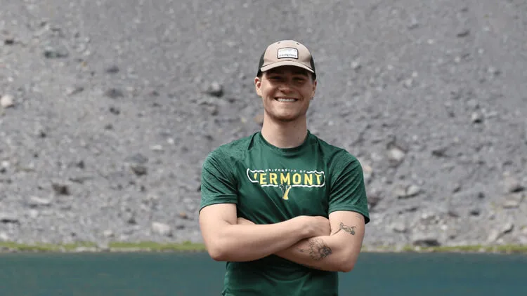 Smiling person wearing a green University of Vermont shirt and tan cap.