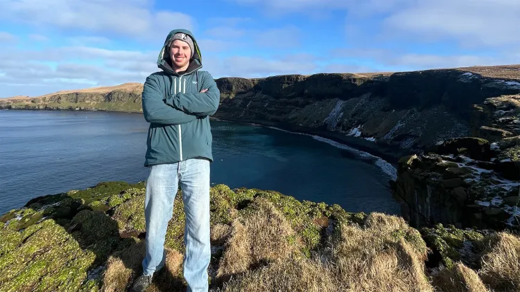 Smiling young man in blue hoodie standing on rocky outcrop with cliffs and sea in backgroun