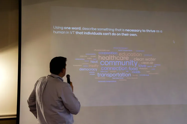 A person stands with their back to the camera, holding a microphone, looking at a projected image of words answering the question: "what does a human need to thrive in Vermont"