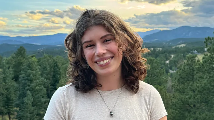 Close-up of smiling young woman with brown hair and white shirt standing with forest and mountains in background 