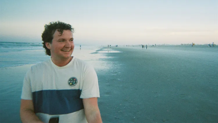 Smiling young man in white and blue t-shirt on beach by the ocean