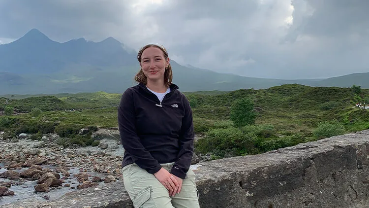 Smiling person sitting on a stone wall in front of a green mountain landscape.