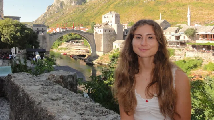 Young woman with long brown hair leaning against stone wall with small village in background