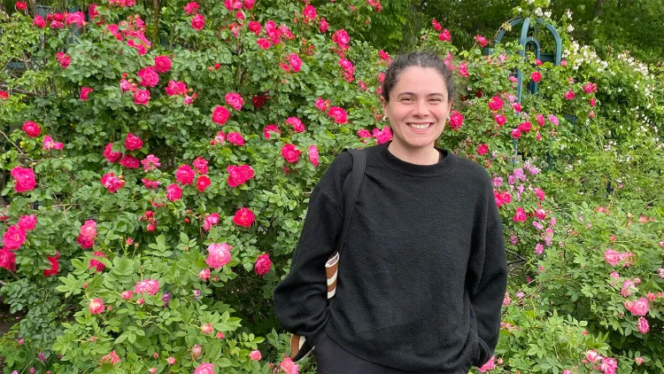 A person smiling while standing outdoors in front of a large, vibrant bush filled with blooming pink roses.