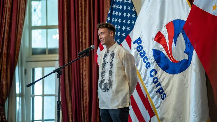 Young man speaking into microphone while standing in front of red, white, and blue Peace Corps flag