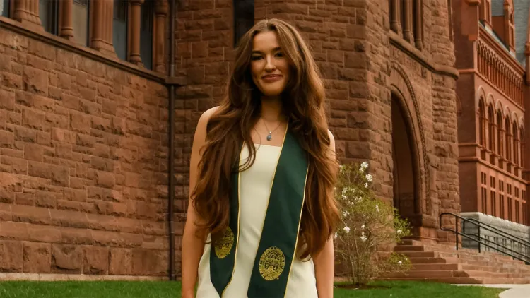 Young woman in white dress and green graduation sash standing in front of red stone academic building