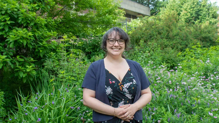 Smiling person standing in a lush garden with flowering plants and trees.