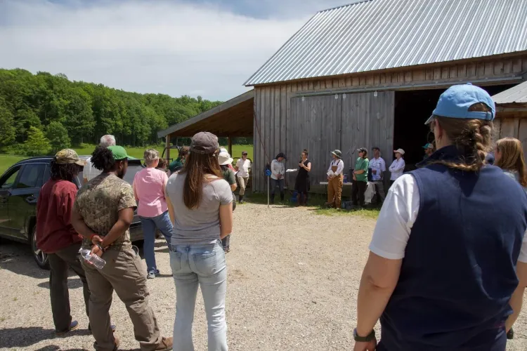 A group of people stand in a large circle outside a barn on a sunny day.