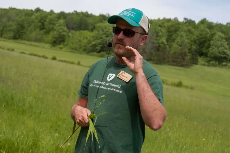 A man in a green shirt, green and white hat and wearing a headset microphone gestures and holds stalks of grass. 