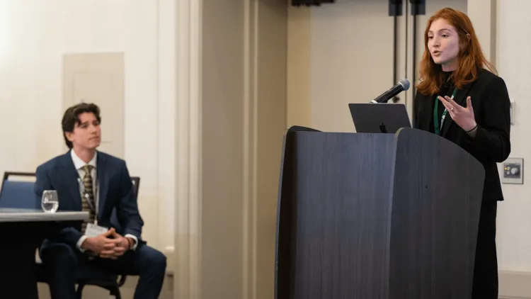a person presenting at a podium as a person seated at a table looks on