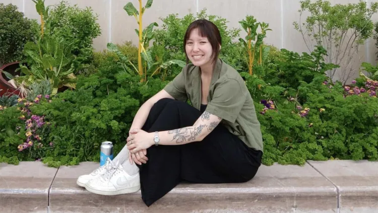 Smiling young woman sitting on stone ledge in front of green bushes 