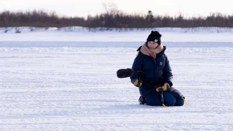 A person in a winter coat kneeling in a snowy landscape while holding a microphone out to capture field audio.