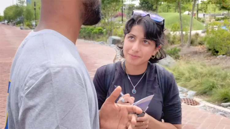 A person holding a notepad and pen looks up attentively at another person during a conversation outdoors.