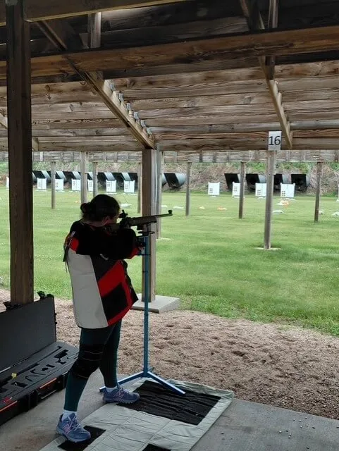 A young person wearing a jacket, spandex and sneakers is seen aiming a long gun at targets from underneath a sheltering overhand. 